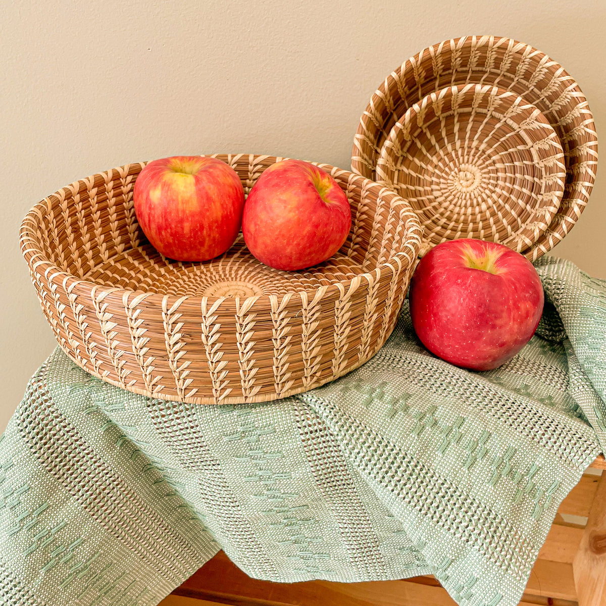 Large Nesting Basket holding two apples, with Medium and Small Nesting Baskets nested alongside it, with one apple in front.  Styled over Sage Calado Table Runner.